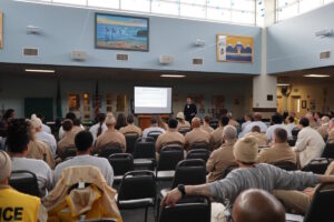  a light filled room with people sitting in chairs arranged in rows watching a man give speak next to a projected image
