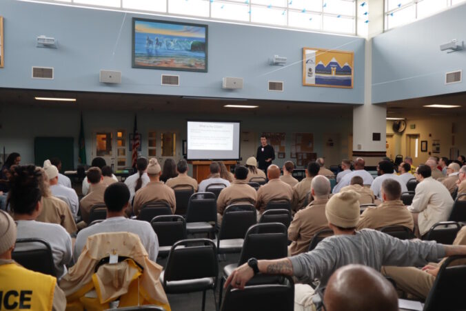 a light filled room with people sitting in chairs arranged in rows watching a man give speak next to a projected image