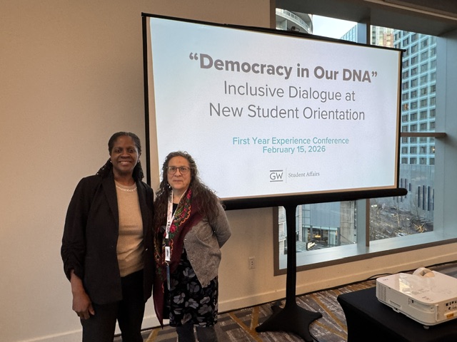 Two women standing side by side near a screen with the title "Democracy in Our DNA" Inclusive Dialogue at New Student Orientation